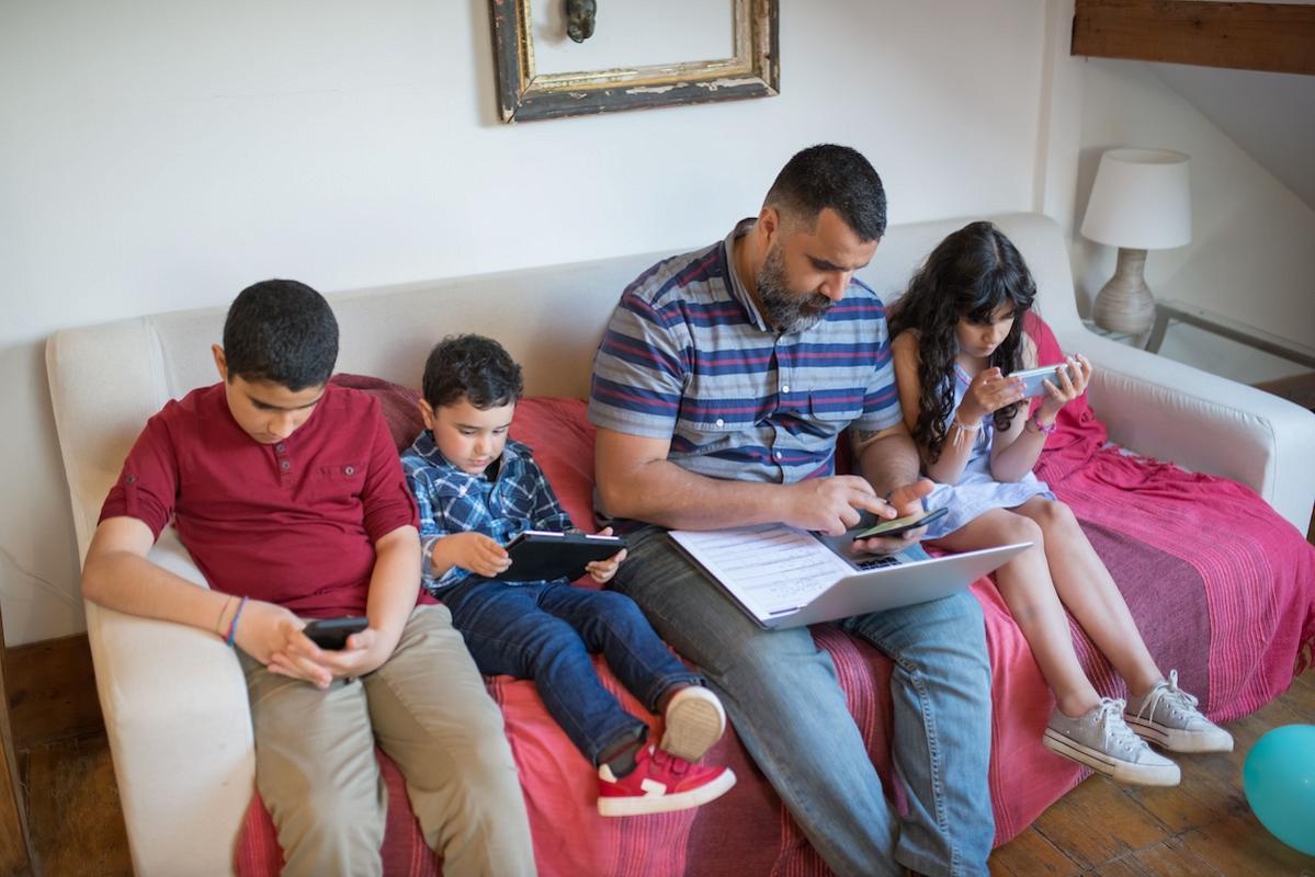 Father and children using gadgets on a sofa, balancing work and leisure.